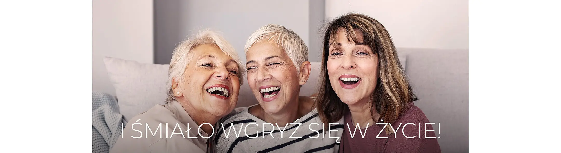 Trzy uśmiechnięte kobiety w różnym wieku siedzą blisko siebie, wyrażając radość i zadowolenie. Na tle jasnego wnętrza widać miłą atmosferę. Na dole napis: „I śmiało wgryź się w życie!”