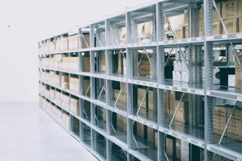 Obraz przedstawia magazyn z metalowymi regałami. Na regałach znajdują się równo poukładane kartony i pudełka, które mogą zawierać różne produkty. Ściana magazynu jest jasna, a podłoga gładka i czysta.