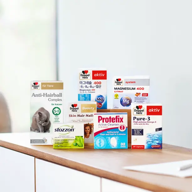 Na stole znajdują się różne produkty zdrowotne marki Doppelherz, w tym suplementy diety takie jak "Anti-Hairball Complex", "Magnesium 400" oraz "Skin-Hair-Nail". Każde opakowanie ma wyraźne napisy i różne kolory, a w centrum stoi mały kotek na opakowaniu.