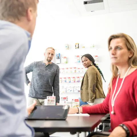 Four people are engaged in a discussion in a well-lit office. One person sits at a table with a tablet, while the others stand and lean in, looking toward the seated individual. A display of products is visible on the wall behind them.