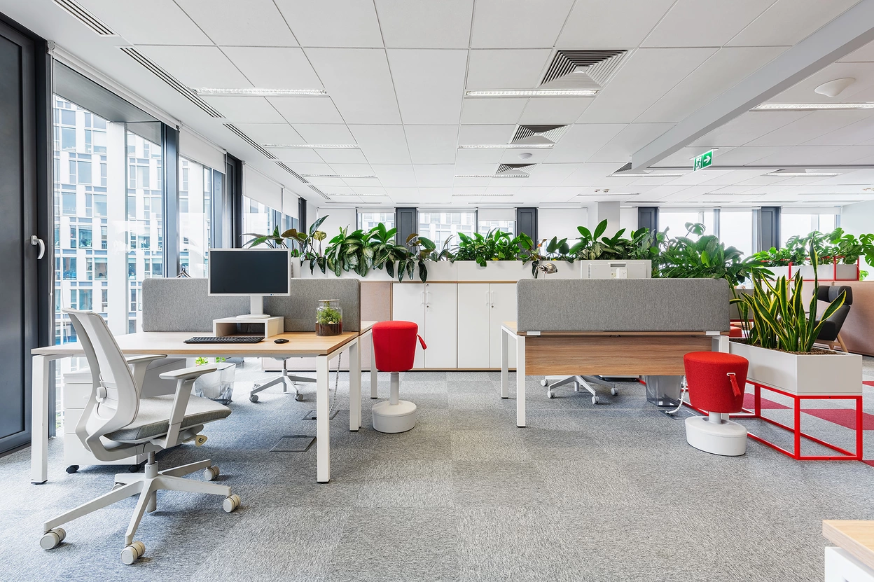 Nowoczesne biuro z dużymi oknami, jasno oświetlone, z roślinami na tle. W centralnej części widoczny drewniany stół z komputerem i ergonomiczne krzesło. Obok niewielki czerwony taboret oraz szafki na dokumenty. Przestrzeń sprzyja pracy i relaksowi.