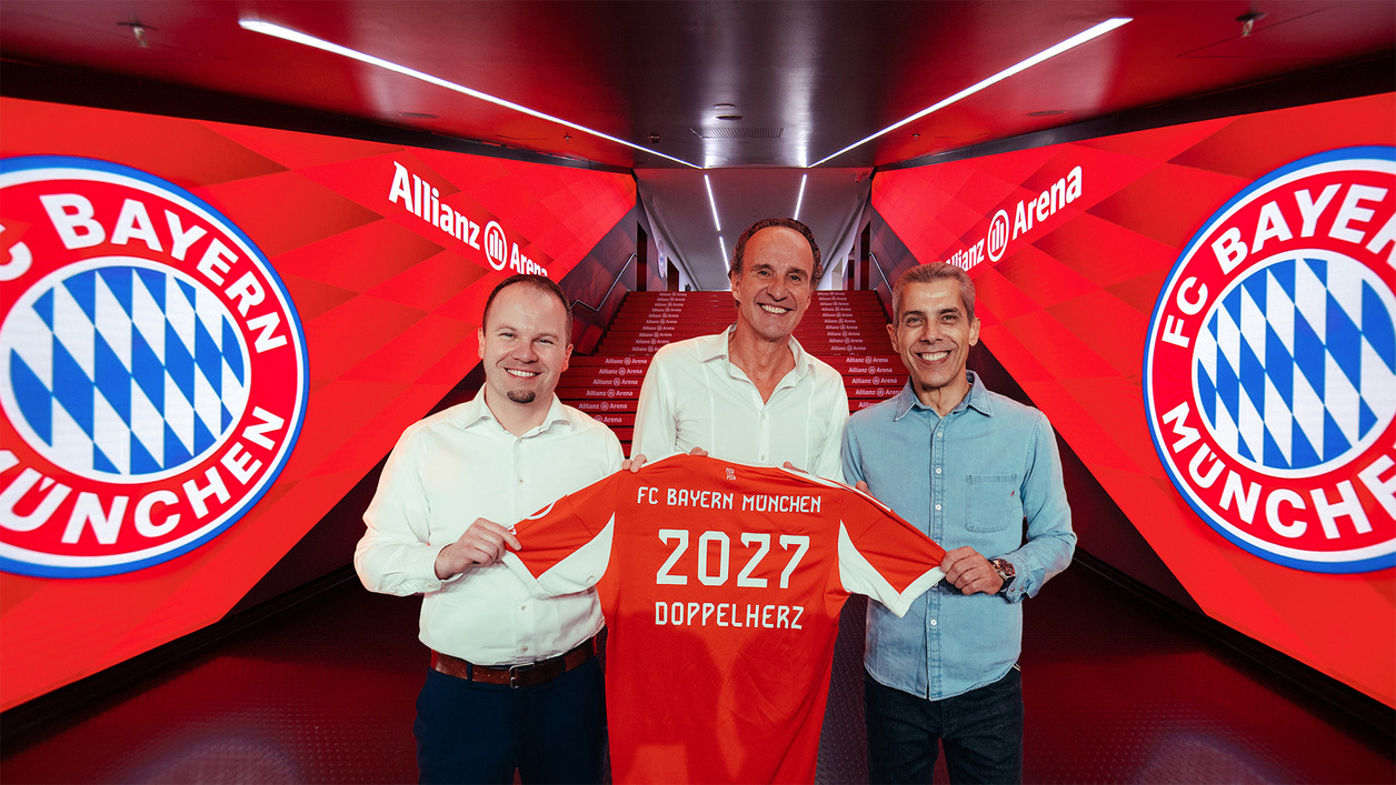 Trzech mężczyzn stoi w tunelu Allianz Areny, trzymając piłkarską koszulkę FC Bayern Monachium z numerem 2027. Na tle widać logo drużyny oraz czerwone i białe dekoracje. Uśmiechają się, wyrażając radość z podpisania umowy.